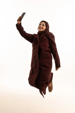 توثيق اللحظات الجميلة، التقاط صور سيلفي باستخدام الهاتف المحمول،بورتريه لشابة عربية خليجية سعودية محجبة ترتدي عباءة تقفز في الهواء بيدها الجوال بتعابير السرور والسعادة، استخدام جهاز تقني حديث، الجسم بالطول الكامل، خلفية بيضاء