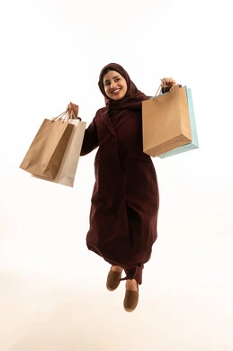 تسوق و شراء الهدايا من المراكز التجارية السعودية,بورتريه لشابة عربية خليجية سعودية محجبة ترتدي عباءة تقفز في الهواء تحمل بيدها مجموعة من حقائب التسوق، شراء الاحتياجات من المول، الجسم بالطول الكامل، خلفية بيضاء