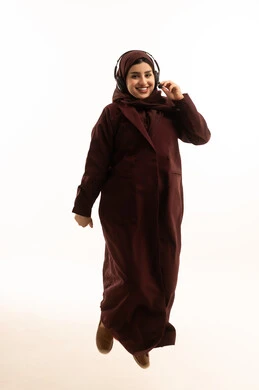 التسلية والترفيه بإستخدام تقنية الهيدفون، بورتريه لشابة عربية خليجية سعودية محجبة ترتدي عباءة تقفز في الهواء تستمتع بسماع الأغاني والموسيقى والصوتيات عبر سماعات الرأس الصوتية، الجسم بالطول الكامل، خلفية بيضاء