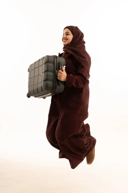 قضاء رحلة صيفية ترفيهية ممتعة، الاستعداد للسفر في جولة سياحية، بورتريه من الجانب لشابة عربية خليجية سعودية محجبة ترتدي عباءة تقفز في الهواء و تحمل حقيبة السفر, النظر بايماءات السعادة و السرور، الجسم بالطول الكامل، خلفية بيضاء