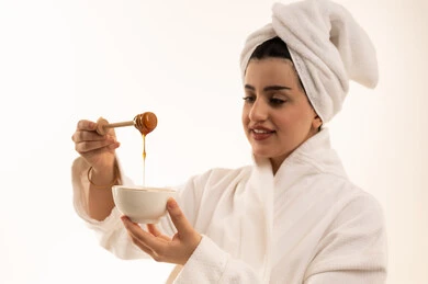 Using honey and natural products for skincare, following a healthy diet, nourishing vitamins for the body, a locally made Saudi product, a close-up portrait of a Saudi Gulf Arab woman wearing a bathrobe and a white towel holding a wooden spoon dipped in honey, full-body image, white background.