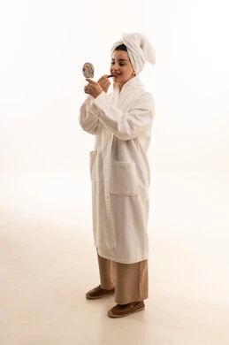 دورات تعليمية لمكياج تتوريال، مفهوم الإهتمام بالجمال والبشرة والمظهر الخارجي, مستحضرات التجميل النسائية، بورتريه لإمرأة عربية خليجية سعودية ترتدي لامرأة عربية خليجية سعودية ترتدي روب استحمام أبيض و منشفة بيضاء تحمل مرآة تقوم بوضع احمر الشفاه،الجسم بالطول الكامل، خلفية بيضاء
