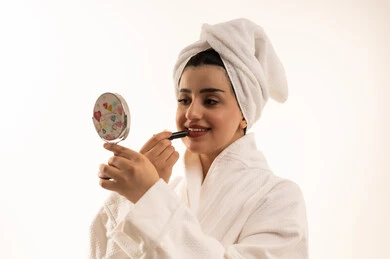 مستحضرات التجميل النسائية، دورات تعليمية لمكياج تتوريال، مفهوم الإهتمام بالجمال والبشرة والمظهر الخارجي، بورتريه مقرب لإمرأة عربية خليجية سعودية ترتدي لامرأة عربية خليجية سعودية ترتدي روب استحمام أبيض و منشفة بيضاء تحمل مرآة تقوم بوضع احمر الشفاه، خلفية بيضاء
