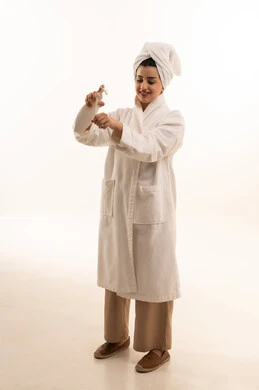 Skincare through creams and products, taking care of the external appearance, a portrait of a Saudi Gulf Arab woman wearing a white bathrobe and a white towel holding a moisturizing cream jar in her hand, full-body image, white background.