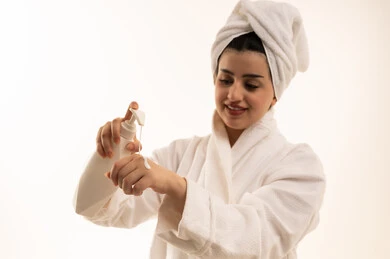 Skincare through cosmetics, attention to appearance, a close-up portrait of a Saudi Gulf Arab woman wearing a white bathrobe and a white towel, holding a moisturizing cream jar in her hand, with a white background.