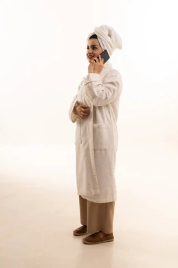 قضاء اوقات ممتعة بتبادل الاخبار والأحاديث، التحدث عبر الهاتف المحمول، بورتريه لامرأة عربية خليجية سعودية ترتدي روب استحمام أبيض تقوم بإجرء مكالمة هاتفية، محادثات ودردشات مع الاصدقاء، صورة الجسم بالكامل، خلفية بيضاء
