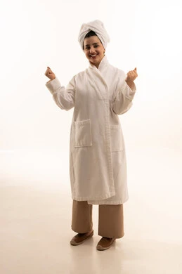 النظر الى الكاميرا بتعابير الفوز والكسب، ايماءات الفرح للانتهاء من انجاز شيء معين، مفهوم النجاح و تحقيق الاهداف، بورتريه لامرأة عربية خليجية سعودية ترتدي روب استحمام أبيض و منشفة ترفع يديها للأعلى بحركات التشجيع والحماس، الجسم بالطول الكامل، خلفية بيضاء