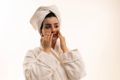 استخدام كريمات ومكونات تجميلية، مفهوم العناية بنضارة وتحسين البشرة، الاهتمام بالمظهر الخارجي، بورتريه مقرب لامرأة عربية خليجية سعودية ترتدي روب استحمام أبيض و منشفة بيضاء تضع كريم الترطيب، خلفية بيضاء