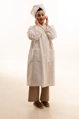 الاهتمام بالمظهر الخارجي، مفهوم العناية بنضارة وتحسين البشرة، بورتريه لامرأة عربية خليجية سعودية ترتدي روب استحمام أبيض و منشفة بيضاء تضع كريم الترطيب، استخدام كريمات ومكونات تجميلية، الجسم بالطول الكامل، خلفية بيضاء