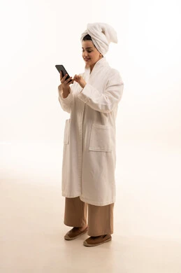 استخدام الأجهزة التقنية الحديثة ,الدردشة مع الأصدقاء و الأهل, تصفح وسائل التواصل الاجتماعي, بورتريه لامرأة عربية خليجية سعودية ترتدي روب استحمام أبيض و منشفة تقوم باستعمال الهاتف المحمول,  الجسم بالطول الكامل، خلفية بيضاء