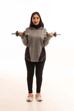 بورتريه لفتاة عربية خليجية سعودية ترتدي الملابس الرياضية، ممارسة التمارين الرياضية مع الدمبل، نشاط بدني للمحافظة على الصحة واللياقة الجسدية، كمال الأجسام، أسلوب حياة صحي، بناء عضل، تمارين رفع الأثقال (دمبلز)، رفع الأوزان الحديدية الثقيلة، صورة بالطول الكامل، خلفية بيضاء