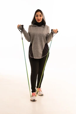 نشاط بدني لتعزيز اللياقة والصحة الجسدية،  ممارسة التمارين الرياضية باستخدام الحبال المطاطية، بورتريه لشابة عربية خليجية سعودية ترتدي زيًا رياضيًا تمارس تمارين المقاومة لشد وتقوية العضلات، مفهوم الرشاقة والنشاط البدني، أسلوب حياة صحي ومفيد، خلفية بيضاء.