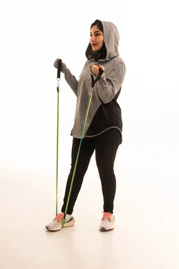 ممارسة التمارين الرياضية باستخدام الحبال المطاطية، نشاط بدني لتعزيز اللياقة والصحة الجسدية، بورتريه لشابة عربية خليجية سعودية ترتدي زيًا رياضيًا تمارس تمارين المقاومة لشد وتقوية العضلات، مفهوم الرشاقة والنشاط البدني، أسلوب حياة صحي ومفيد، خلفية بيضاء.