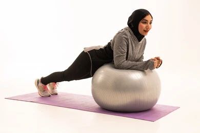 Physical activity to maintain health and physical fitness, an Arab Gulf Saudi girl wearing sportswear, exercising with a fitness ball, a healthy lifestyle, practicing Pilates ball and balance exercises, white background.