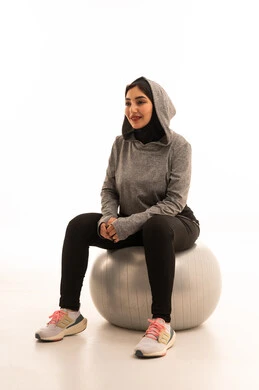 Exercising with a fitness ball, an Arab Gulf Saudi girl wearing sportswear, physical activity to maintain health and physical fitness, a healthy lifestyle, practicing Pilates ball and balance exercises, white background.