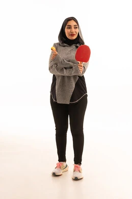 ممارسة رياضات وانشطة بدنية ممتعة، اللعب بسرعة وتركيز، بورتريه  لشابة عربية خليجية سعودية ترتدي ملابس رياضية تحمل بيدها مضرب تنس الطاولة، المحافظة على الرياضة والرشاقة الجسدية، خلفية بيضاء