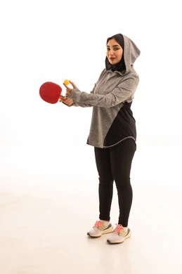 المحافظة على الرياضة والرشاقة الجسدية، اللعب بسرعة وتركيز، ممارسة رياضات وانشطة بدنية ممتعة، بورتريه  لشابة عربية خليجية سعودية ترتدي ملابس رياضية تحمل بيدها مضرب تنس الطاولة،خلفية بيضاء