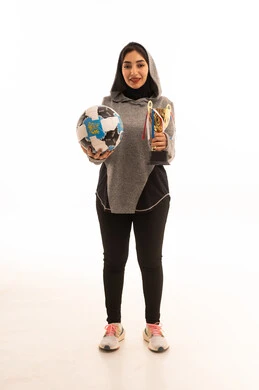 النظر إلى الكاميرا بإيماءات السعادة بالفوز والنصر، احتراف الرياضات الجماعية، المحافظة على نمط حياة صحي، بورتريه مقرب للاعبة عربية خليجية سعودية محجبة مبتسمة ترتدي الملابس الرياضية تحمل كرة القدم وكأس البطولة، خلفية بيضاء