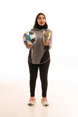 النظر إلى الكاميرا بإيماءات السعادة بالفوز والنصر، احتراف الرياضات الجماعية، المحافظة على نمط حياة صحي، بورتريه مقرب للاعبة عربية خليجية سعودية محجبة مبتسمة ترتدي الملابس الرياضية تحمل كرة القدم وكأس البطولة، خلفية بيضاء