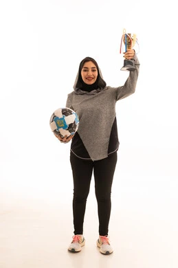 المحافظة على نمط حياة صحي، النظر إلى الكاميرا بإيماءات السعادة بالفوز والنصر، احتراف الرياضات الجماعية، بورتريه مقرب للاعبة عربية خليجية سعودية محجبة مبتسمة ترتدي الملابس الرياضية تحمل كرة القدم وكأس البطولة، خلفية بيضاء