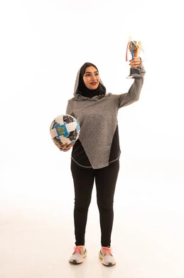 احتراف الرياضات الجماعية، النظر إلى الكاميرا بإيماءات السعادة بالفوز والنصر، المحافظة على نمط حياة صحي، بورتريه مقرب للاعبة عربية خليجية سعودية محجبة مبتسمة ترتدي الملابس الرياضية تحمل كرة القدم وكأس البطولة، خلفية بيضاء