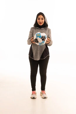 بورتريه  للاعبة عربية خليجية سعودية محجبة مبتسمة ترتدي الملابس الرياضية تحمل كرة القدم وكأس البطولة، النظر إلى الكاميرا بإيماءات السعادة بالفوز والنصر، احتراف الرياضات الجماعية، المحافظة على نمط حياة صحي،  خلفية بيضاء