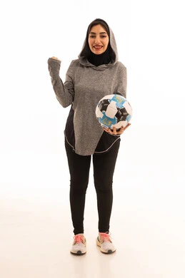 المحافظة على نمط حياة صحي، النظر إلى الكاميرا بإيماءات السعادة بالفوز والنصر، احتراف الرياضات الجماعية، بورتريه مقرب للاعبة عربية خليجية سعودية محجبة مبتسمة ترتدي الملابس الرياضية تحمل كرة القدم وكأس البطولة، خلفية بيضاء
