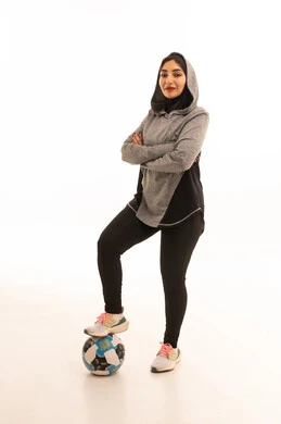 النظر إلى الكاميرا بإيماءات السعادة بالفوز والنصر، احتراف الرياضات الجماعية، المحافظة على نمط حياة صحي، بورتريه مقرب للاعبة عربية خليجية سعودية محجبة مبتسمة ترتدي الملابس الرياضية تحمل كرة القدم وكأس البطولة، خلفية بيضاء