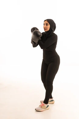 التدرب على الحركات القتالية ،  المحافظة على اللياقة البدنية، زيادة قدرة العضلات على التحمل، بورتريه لفتاة عربية خليجية سعودية ترتدي الملابس الرياضية والقفازات تنظر إلى الكاميرا تتعلم فنون الملاكمة، تعزيز صحة العظام والمفاصل، خلفية بيضاء