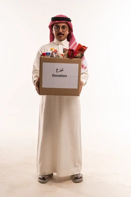 التصدق بالفائض من المستلزمات، جمعية خيرية ومبادرة انسانية، الانفاق والتبرع في سبيل الله للجمعيات السعودية، النظر الى الكاميرا بتعابير السرور، بورتريه لرجل  خليجي عربي سعودي يرتدي الثوب التقليدي والشماغ يحمل بيده صندوق التبرعات ويمسك مواد غذائية، خلفية بيضاء