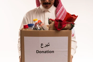 الانفاق والتبرع في سبيل الله للجمعيات السعودية، جمعية خيرية ومبادرة انسانية، التصدق بالفائض من المستلزمات، النظر الى الكاميرا بتعابير السرور، بورتريه مقرب لرجل  خليجي عربي سعودي يرتدي الثوب والشماغ التقليدي يحمل بيده صندوق التبرعات فيه مواد غذائية، خلفية بيضاء