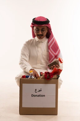 Islamic charitable works in Saudi Arabia, a charitable association and humanitarian initiative, spending and donating for the sake of God to Saudi associations, a portrait of a Gulf Arab Saudi man wearing traditional attire and a ghutrah holding food supplies in his hand, wearing traditional clothing and glasses, with a white background.