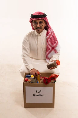 الجلوس على الركبتين والتعرف على صندوق التبرع، جمعية خيرية ومبادرة انسانية، الانفاق والتبرع في سبيل الله للجمعيات السعودية، النظر الى الكاميرا بتعابير السرور، بورتريه  لرجل  خليجي عربي سعودي يرتدي الثوب التقليدي والشماغ يحمل بيده مواد غذائية، خلفية بيضاء