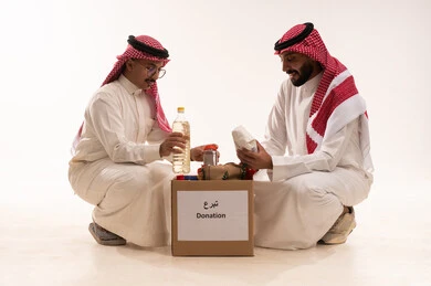 التأكد من صلاحية المواد الغذائية قبل التوزيع، بورتريه لرجلان خليجيان عربيان سعوديان يقومان بتجهيز صندوق المواد الغذائية، التبرع لمشروع السلة الغذائية، توفير الطعام للمحتاجين، مبادرات وتبرعات انسانية خيرية، جمعيات اسلامية في السعودية، شهر رمضان، خلفية بيضاء