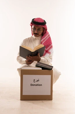 تعابير الالاندماج والتركيز، الانفاق والتبرع في سبيل الله للجمعيات، جمعية خيرية ومبادرة انسانية، التصدق بالفائض من المستلزمات، الأعمال الصالحة في أقات فضيلة، بورتريه مقرب لرجل  خليجي عربي سعودي يرتدي الثوب التقليدي والشماغ يحمل بيده كتاب ويقرأه، خلفية بيضاء