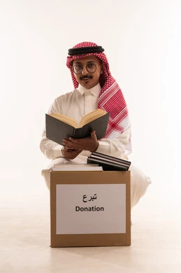 التبرع  والانفاق في سبيل الله للجمعيات، تعابير الاندماج والتركيز، جمعية خيرية ومبادرة انسانية، التصدق بالفائض من المستلزمات، الأعمال الصالحة في أقات فضيلة، بورتريه مقرب لرجل  خليجي عربي سعودي يرتدي الثوب التقليدي والشماغ يحمل بيده كتاب ويقرأه، خلفية بيضاء