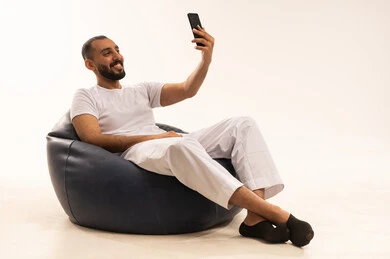 Taking commemorative photos, a selfie, using modern technology devices, making video calls over the internet, a portrait of an Arab Saudi Gulf man wearing white pants and a t-shirt sitting on a comfortable chair holding a mobile phone, the concept of masculinity and self-confidence, white background.