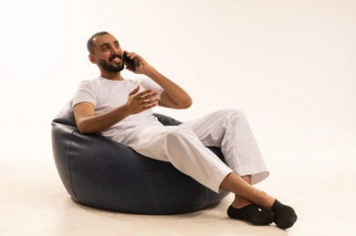 Making a phone call using modern devices, using modern technology devices, making video calls over the internet, a portrait of an Arab Saudi Gulf man wearing white pants and a t-shirt sitting on a comfortable chair holding a mobile phone, the concept of masculinity and self-confidence, white background.