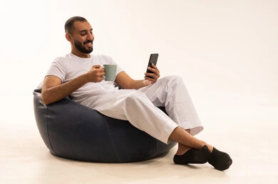 شرب القهوة في وقت تصفح المواقع الاجتماعية ، استخدام اجهزة تقنية حديثة، إجراء مكالمات الفيديو عبر الانترنت، بورتريه  لرجل عربي سعودي خليجي يرتدي سروال وفانيلا باللون أبيض يجلس على كرسي مريح ويحمل جهاز الهاتف المحمول ، مفهوم الرجولة والثقة بالنفس،  خلفية بيضاء