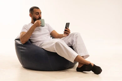 الاستمتاع بشرب المشروبات الساخنة ، استخدام اجهزة تقنية حديثة، تصفح مواقع التواصل، إجراء مكالمات الفيديو عبر الانترنت، بورتريه  لرجل عربي سعودي خليجي يرتدي سروال وفانيلا باللون أبيض يجلس على كرسي مريح ويحمل جهاز الهاتف المحمول ، مفهوم الرجولة والثقة بالنفس،  خلفية بيضاء