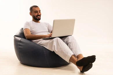 Browsing social media, using modern technology devices, making video calls over the internet, a portrait of an Arab Saudi Gulf man wearing white pants and a t-shirt sitting on a comfortable chair and using a laptop, the concept of masculinity and self-confidence, white background.