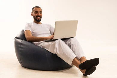 Looking at the camera with gestures of joy and happiness, using modern technological devices, making video calls over the internet, a portrait of an Arab Saudi Gulf man wearing white pants and a t-shirt sitting on a comfortable chair and using a laptop, the concept of masculinity and self-confidence, white background.