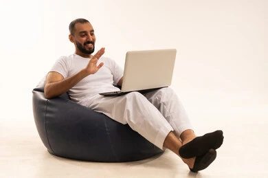 Greeting and peace through the laptop screen, using modern technological devices, making video calls over the internet, a portrait of an Arab Saudi Gulf man wearing white pants and a t-shirt sitting on a comfortable chair and using a laptop, the concept of masculinity and self-confidence, white background.