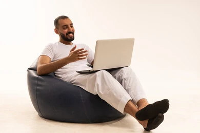 دمج التكنولوجيا مع الحياة العملية، استخدام اجهزة تقنية حديثة، إجراء مكالمات الفيديو عبر الانترنت، بورتريه  لرجل عربي سعودي خليجي يرتدي سروال وفانيلا باللون  أبيض يجلس على كرسي مريح ويستخدم جهاز الحاسب المحمول، مفهوم الرجولة والثقة بالنفس،  خلفية بيضاء