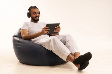 إجراء مكالمات الفيديو عبر الانترنت، العمل و إنجاز المهام عن بعد، استخدام اجهزة تقنية حديثة، بورتريه مقرب لرجل عربي سعودي خليجي يرتدي سروال وفانيلا باللون أبيض يجلس على كرسي مريح ويستخدم جهاز التابلت،يضع على رأسه سماعة هيدفون،مفهوم الرجولة والثقة بالنفس،  خلفية بيضاء