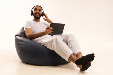 The use of modern technology devices, making video calls over the internet, working and completing tasks remotely, a portrait of an Arab Saudi Gulf man wearing white pants and a t-shirt sitting on a comfortable chair using a tablet, wearing headphones on his head, the concept of masculinity and self-confidence, white background.