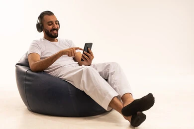 التسوق عبر الإنترنت، باستخدام الأجهزة التكنولوجية الحديثة، تصفح مواقع التواصل الاجتماعي، إجراء مكالمات فيديو عبر الإنترنت، صورة لرجل خليجي سعودي يرتدي سروال وفانيلا باللون الأبيض يجلس على كرسي مريح ويمسك بجهاز محمول ويرتدي سماعات، مفهوم الذكورة والثقة بالنفس، خلفية بيضاء.