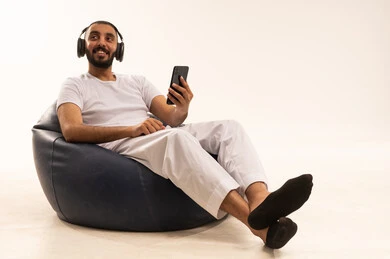Expressions of contemplation and imagination, using modern technological devices, browsing social media, making video calls over the internet, a portrait of an Arab Saudi Gulf man wearing white pants and a t-shirt sitting on a comfortable chair holding a laptop and wearing headphones, the concept of masculinity and self-confidence, white background.