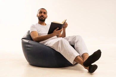 النظر إلى شيء ما، قضاء وقت الفراغ بالتعلم والثقافة، مفهوم محو الامية، تعابير الاندماج والتركيز، هواية حب المطالعة والاستكشاف, بورتريه لرجل عربي خليجي سعودي يرتدي سروال وفانيلا باللون الأبيض يجلس على كرسي مريح يحمل بيده كتاب علمي تثقيفي وينظر الى شيء ما، خلفية بيضاء