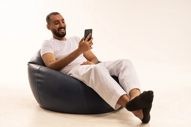 Talking and having fun with friends online, using modern technology devices, making video calls over the internet, a portrait of an Arab Saudi Gulf man wearing white pants and a t-shirt sitting on a comfortable chair holding a mobile phone, the concept of masculinity and self-confidence, white background.